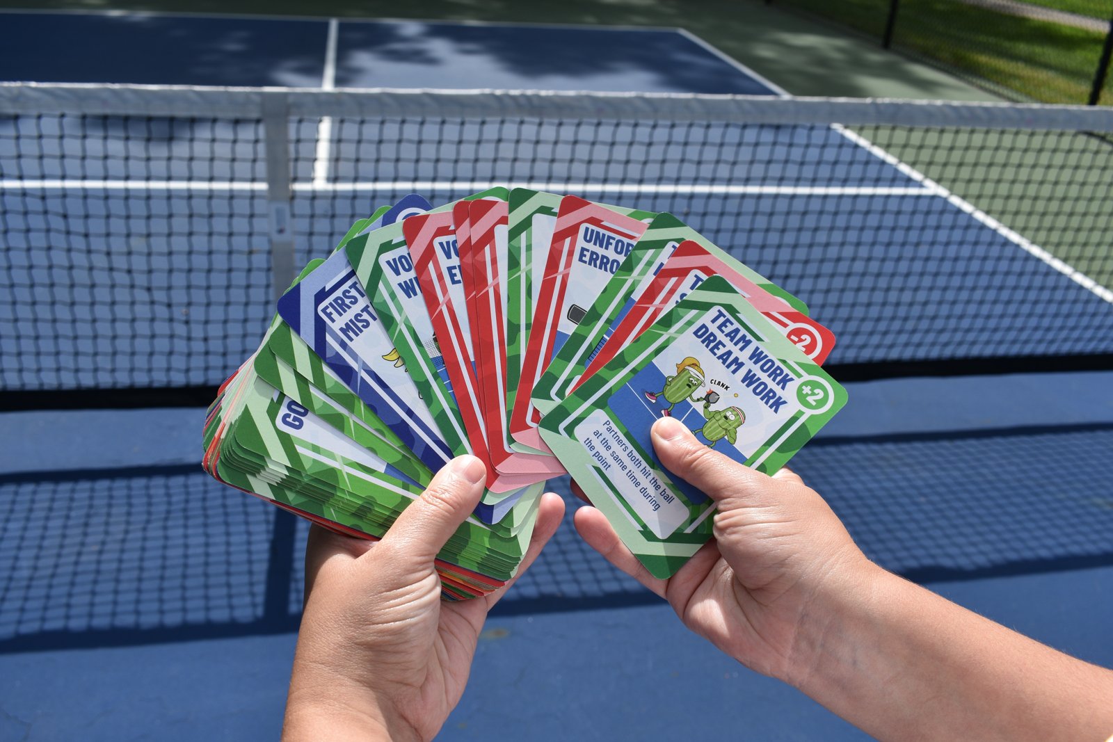 Hands fanning out Dink Deck cards in front of a pickleball net